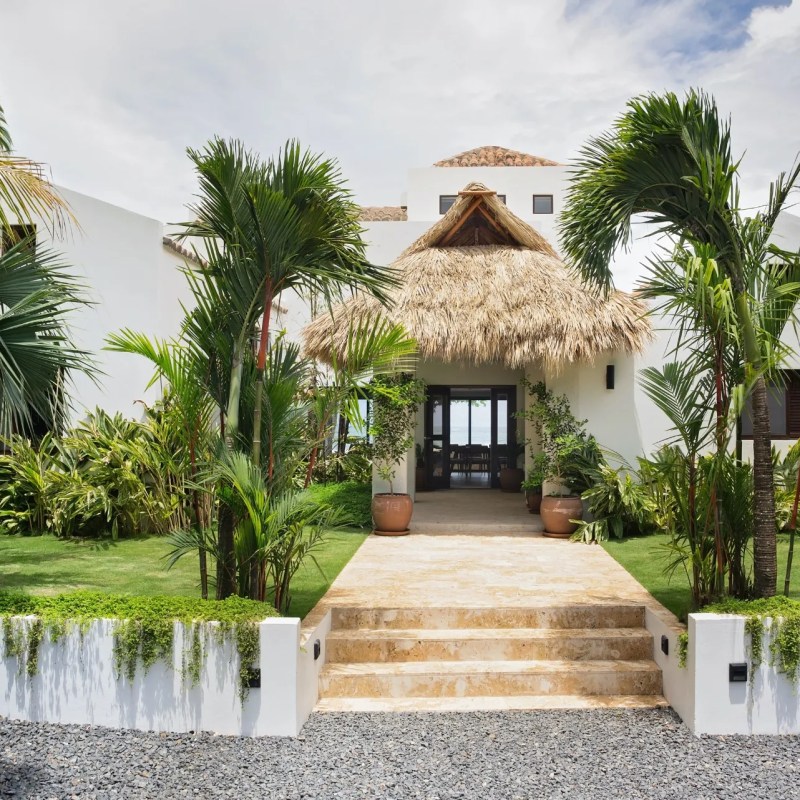 Tropical-style house with thatched roof, surrounded by palm trees and plants.