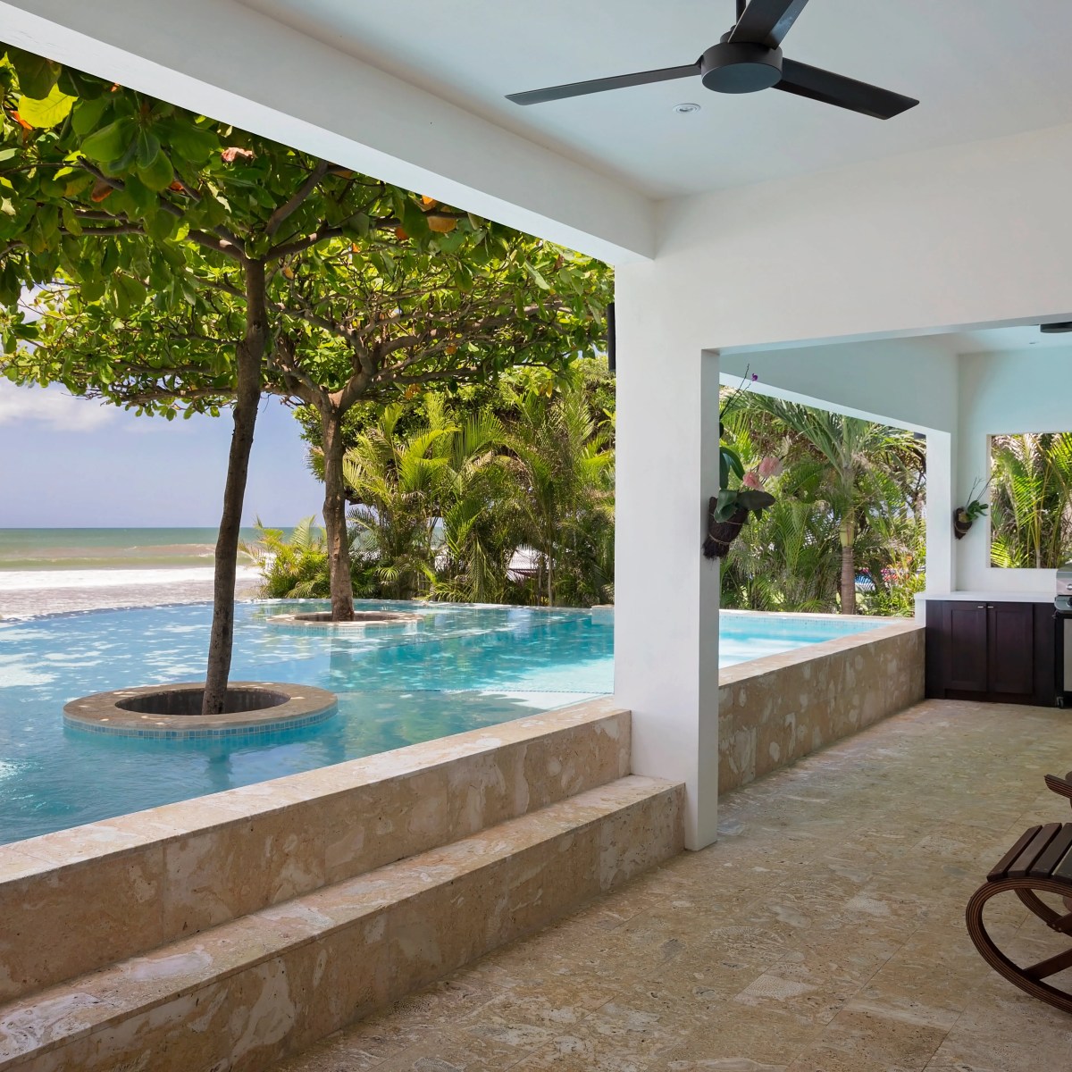 Outdoor patio with rocking chairs, pool, trees, and ocean view under a ceiling fan.