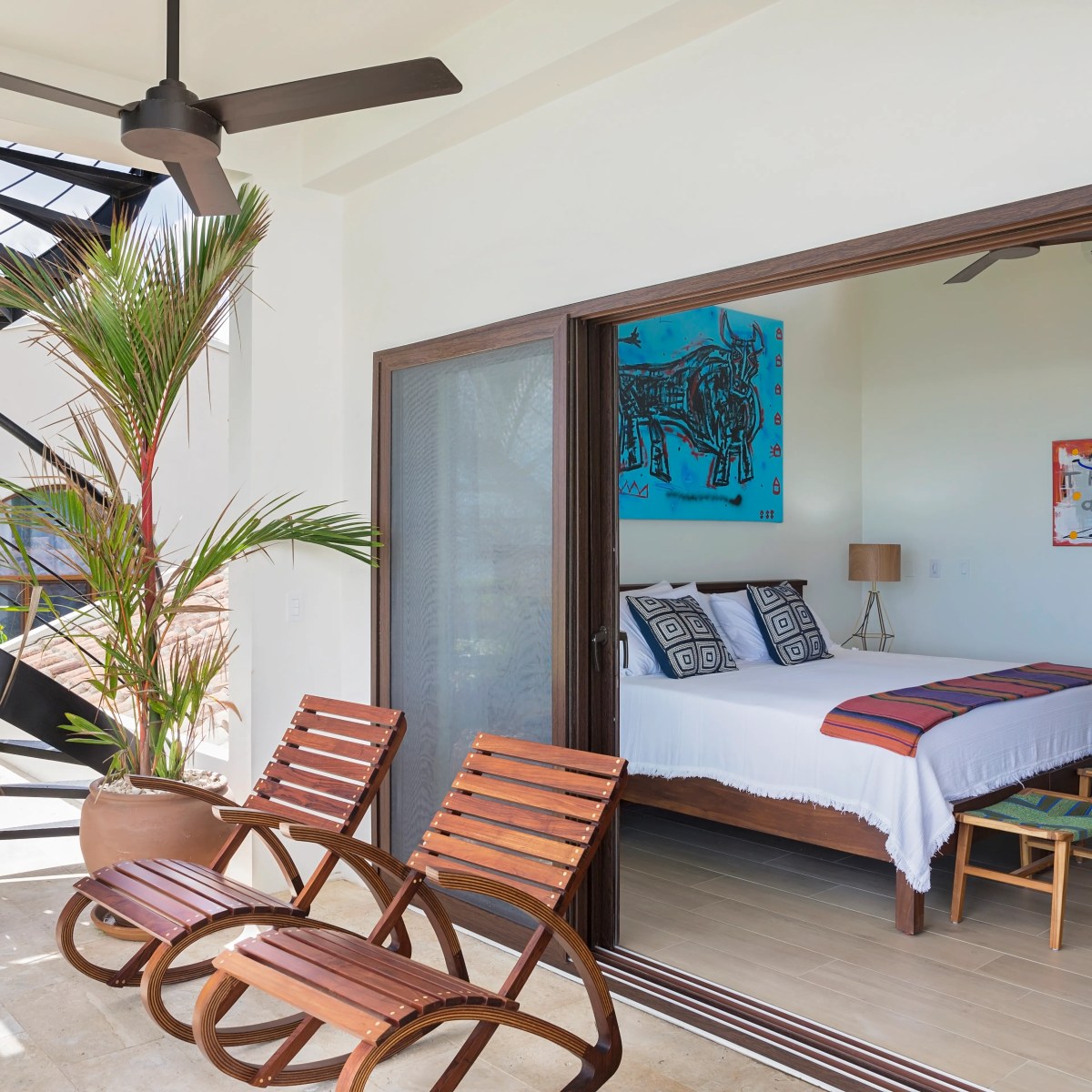 Modern patio with two wooden chairs, a spiral staircase, and view into a bedroom with art on walls.