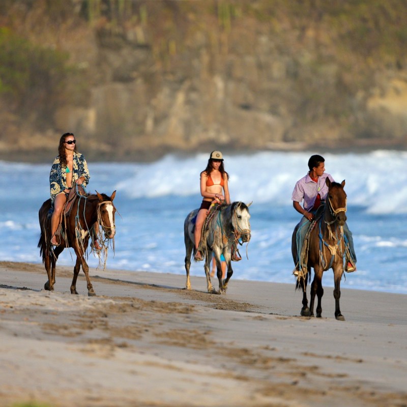 Three people horseback riding on a sandy beach near ocean waves.
