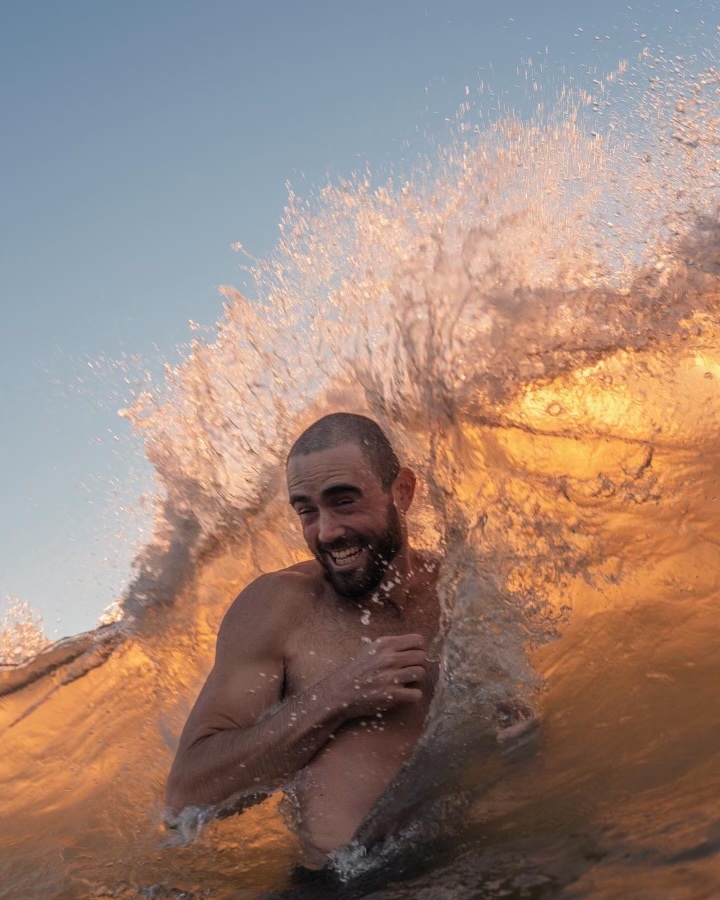 519A68CD-55C2-4C13-9E0A-048B0BB1CFA6 Man with a beard smiles while a wave crashes behind him at sunset.