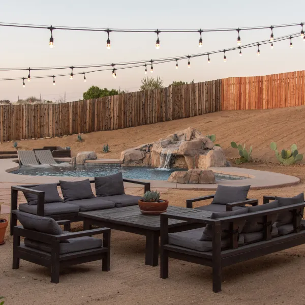 Desert backyard with string lights, seating, pool, waterfall, and wooden fence.