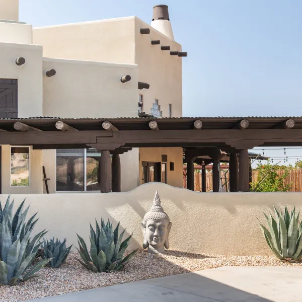 Adobe-style building with wooden beams, agave plants, and a Buddha head sculpture in the garden.