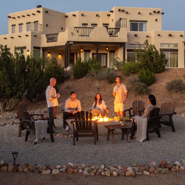 Group relaxing around campfire in front of modern adobe house.