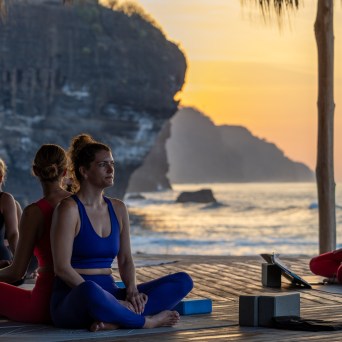Four people practicing yoga on a deck by the ocean at sunset.