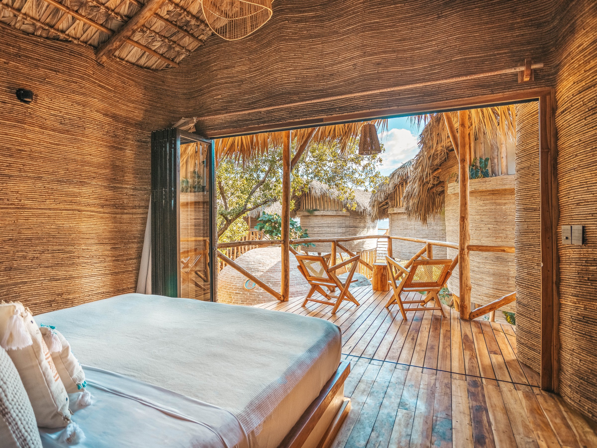 Rustic bedroom with a large bed, open to a wooden deck with chairs and tropical thatched roofs.
