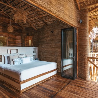 Rustic bedroom with wooden walls, thatched ceiling, and a large bed with decorative pillows.