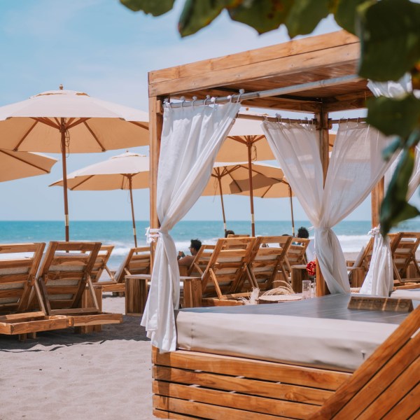 Wooden cabana with white curtains on a sandy beach, surrounded by sun loungers and umbrellas.