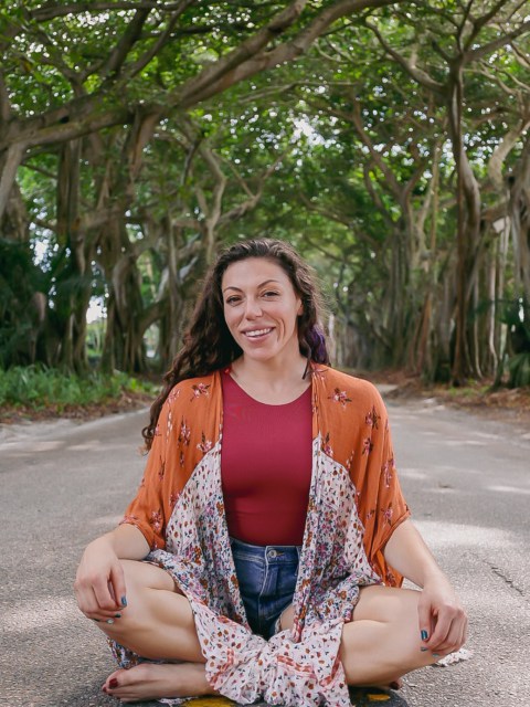 Woman sitting cross-legged on a road under a canopy of trees, smiling at the camera.