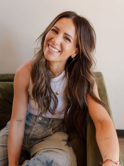 Smiling woman with long hair, wearing a white tank top and jeans, sitting on a green couch.