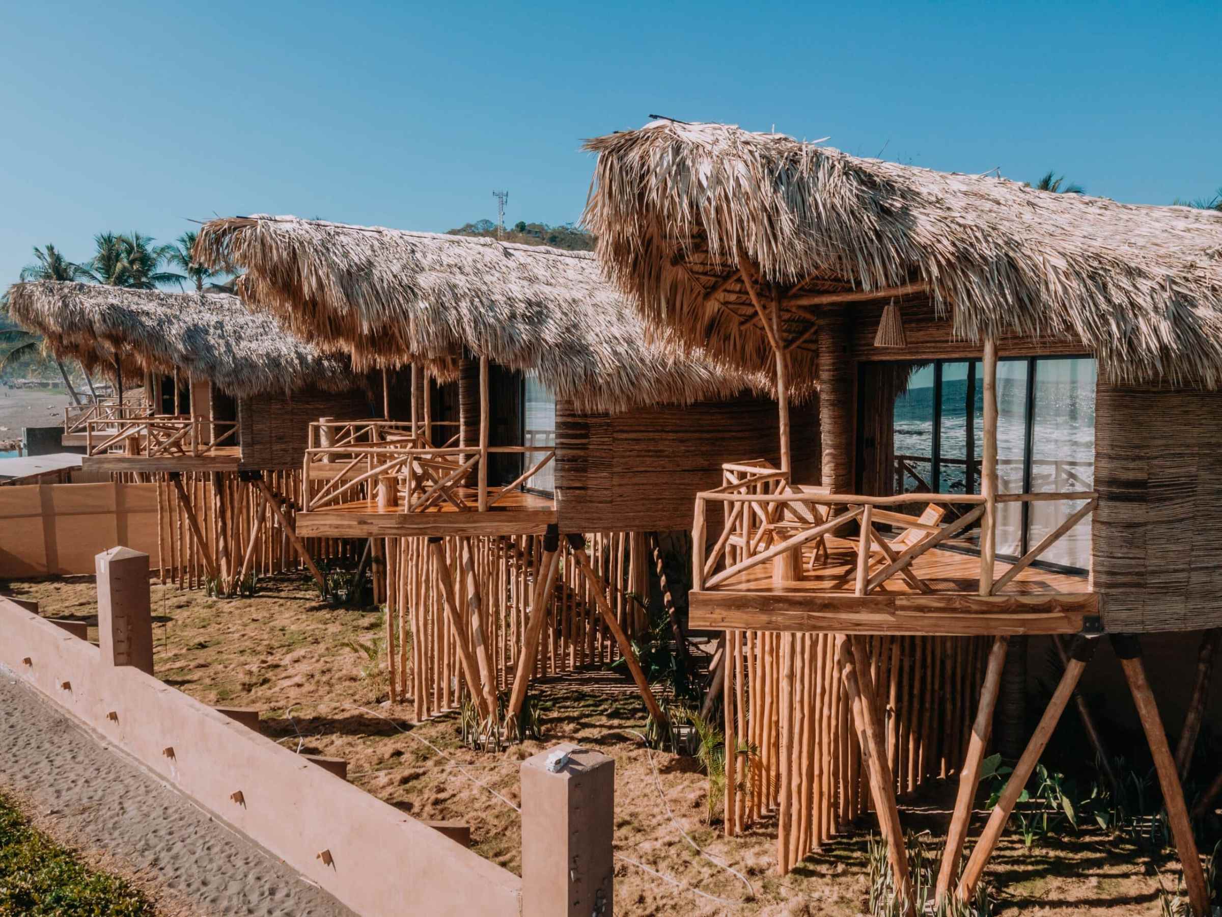 Three thatched-roof bungalows on stilts with bamboo railings, set against a clear blue sky.