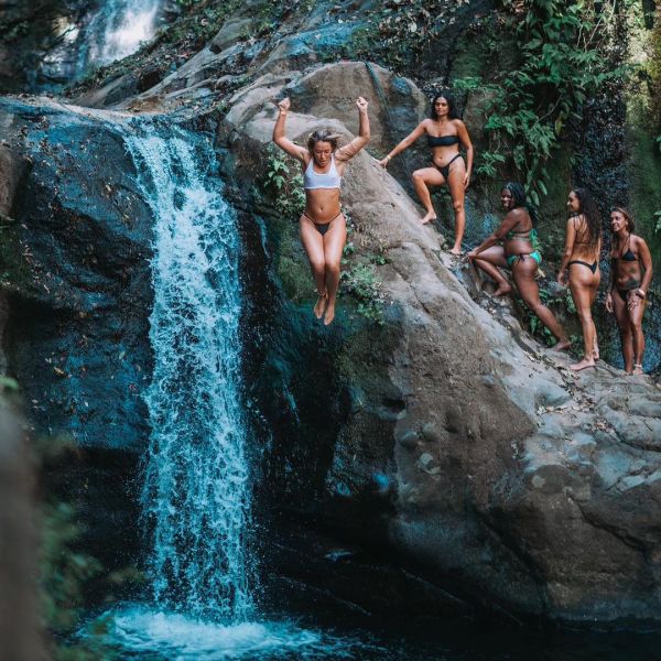 Person jumping off rock near waterfall, group of people watching on rocky ledge.