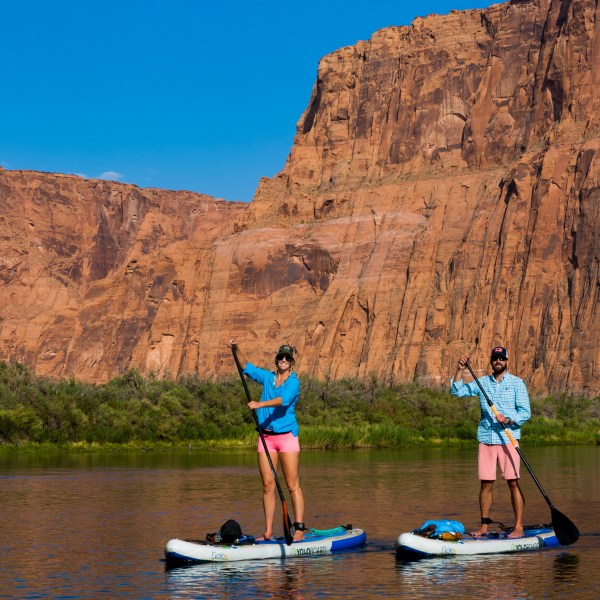Two people paddleboarding on a river, with large red cliffs and clear blue sky in the background.