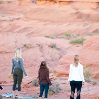 Three people standing on yoga mats in a desert landscape, facing rock formations.