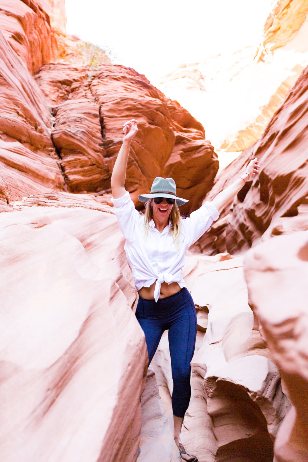 Woman in hat and sunglasses hiking between red rock formations, arms raised, wearing a white shirt and dark pants.