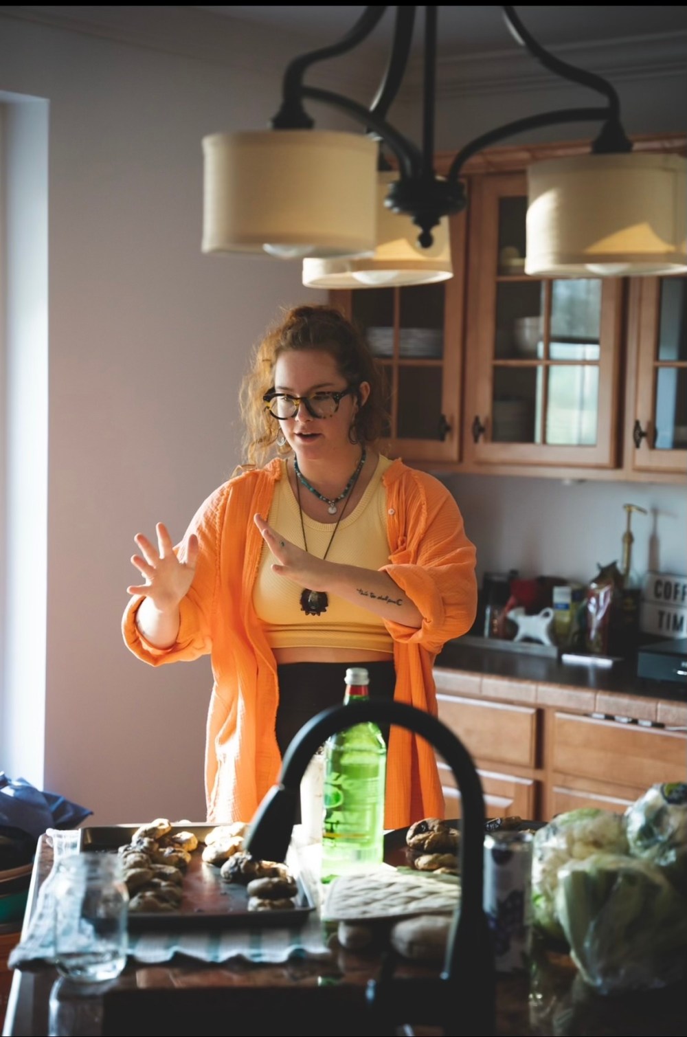 Person in orange shirt gestures while standing in kitchen with cookies and soda on counter.