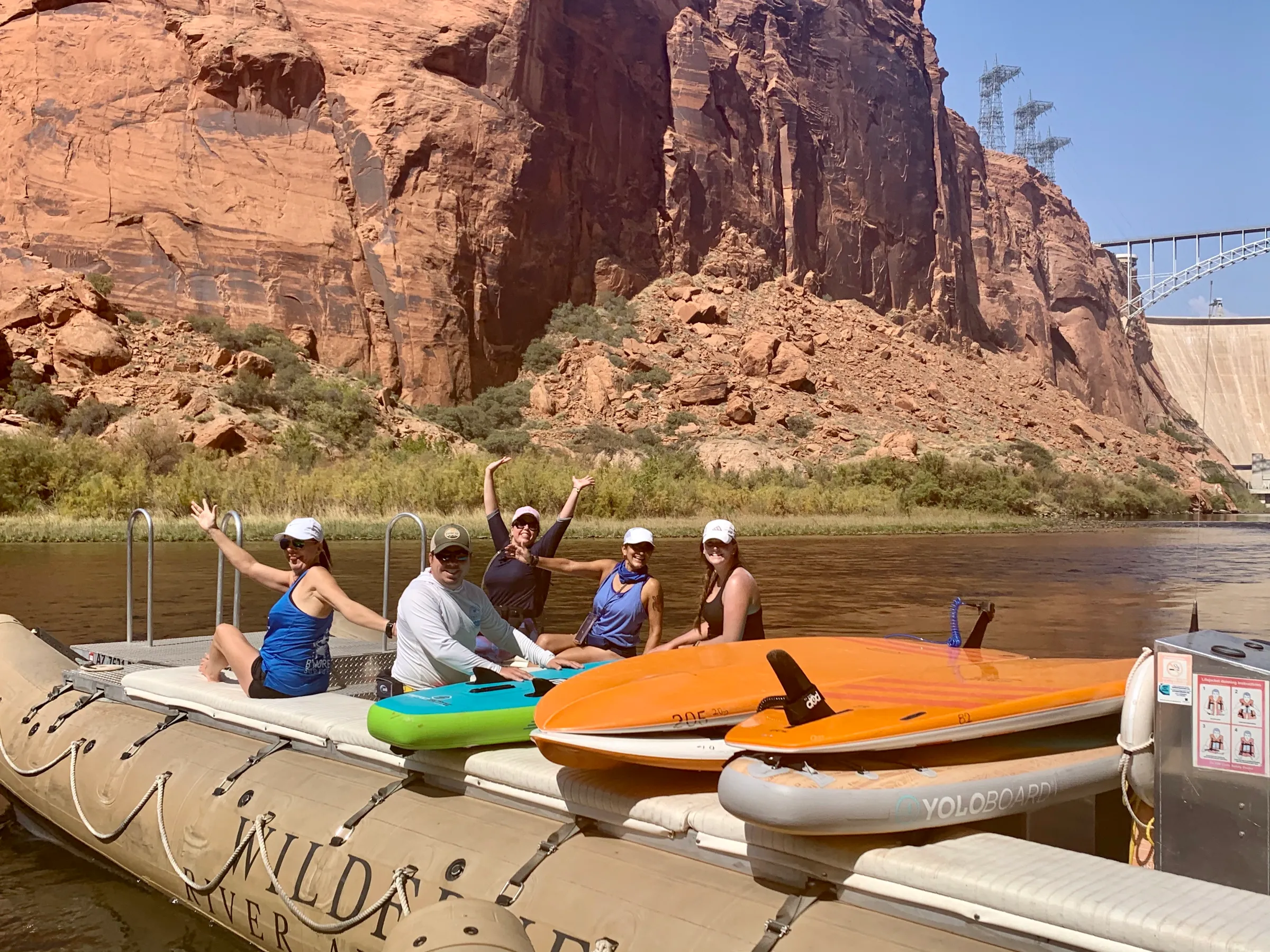 Five people on a boat near a rocky canyon wall with kayaks and paddleboards on a sunny day.