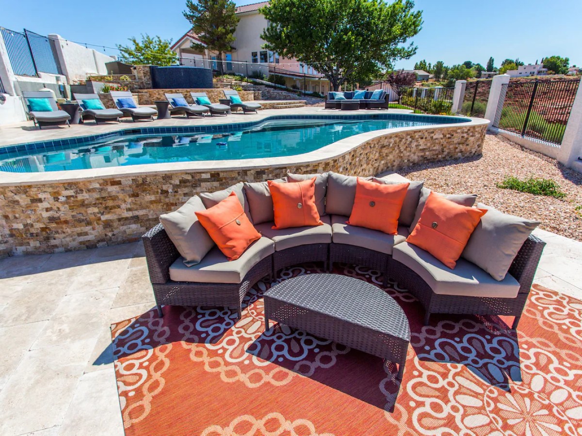 Outdoor pool area with curved sofa, orange cushions, and sun loungers on a sunny patio.