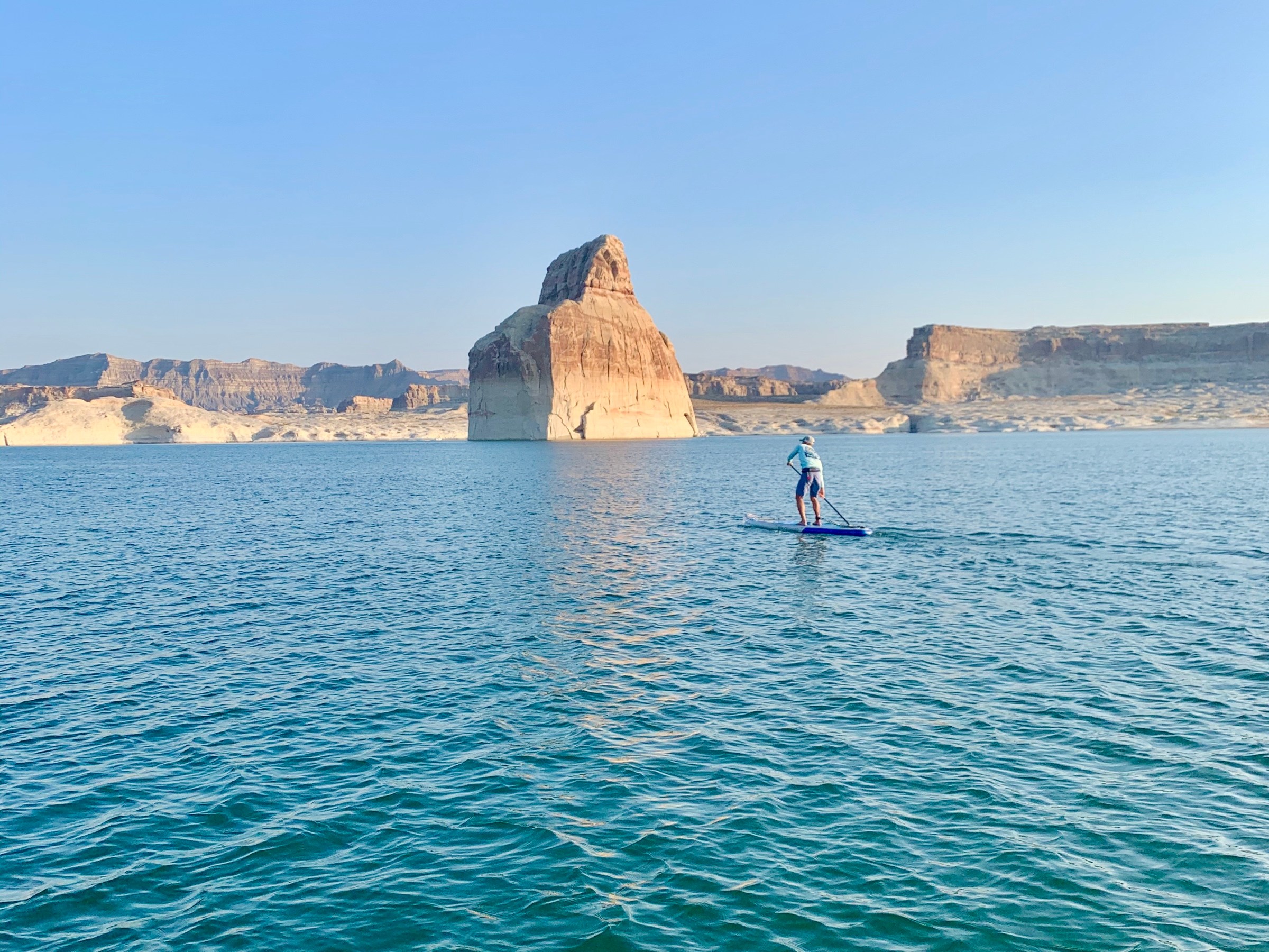 Person paddleboarding on a lake with large rock formation in the background under clear blue sky.