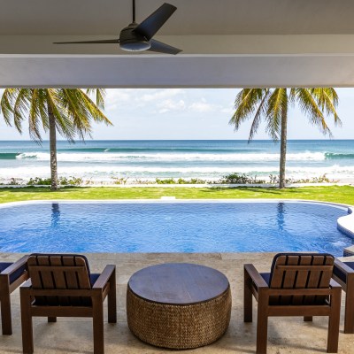 Beachfront view from a patio with chairs facing a pool, palm trees, and ocean waves.