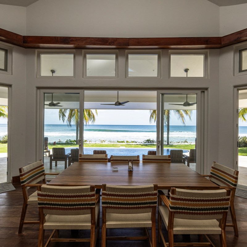 Dining room with wooden table, ocean view through large windows and palm trees outside.