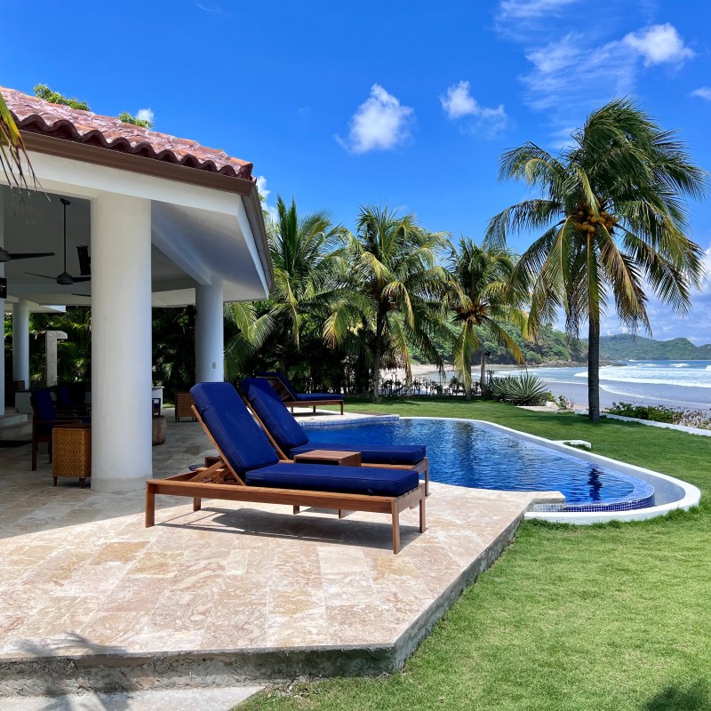 Beachfront villa with pool, lounge chairs, palm trees, and ocean view on a sunny day.
