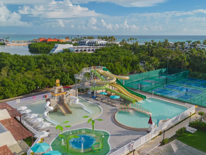 Aerial view of a water park with slides, pools, tennis courts, and lush greenery near the ocean.