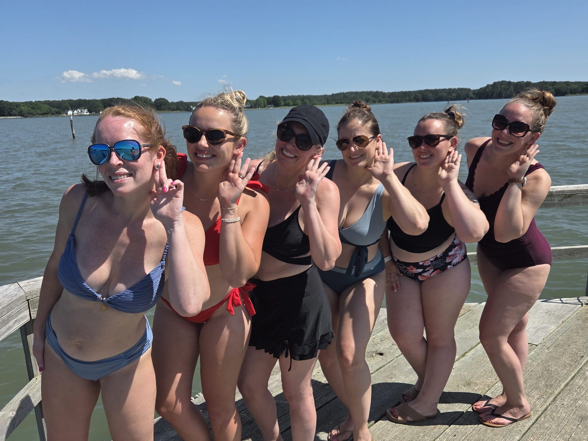 Six women in swimsuits posing on a pier by a lake, all wearing sunglasses, with clear blue sky.