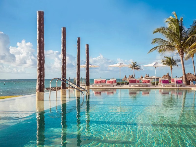 Pool with tall posts, palm trees, loungers, umbrellas, by the ocean under a blue sky.