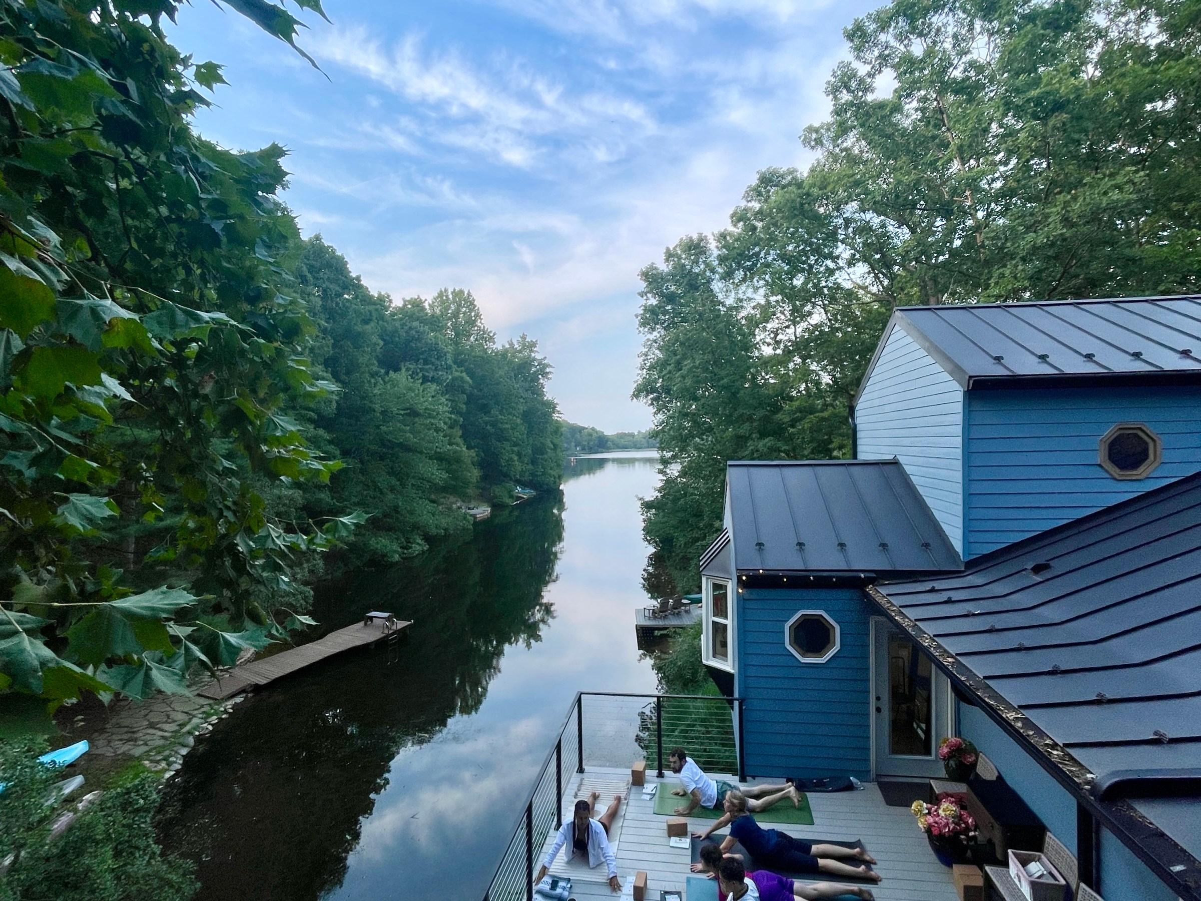Group doing yoga on a deck by a river, with trees and a house visible.