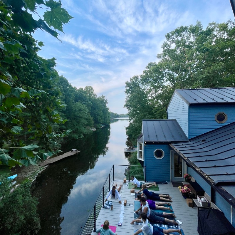 Group doing yoga on a deck by a river, with trees and a house visible.