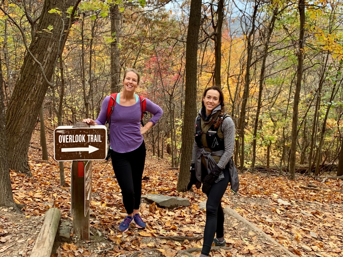 Two women hiking on a forest trail with fall leaves and trees surrounding them.