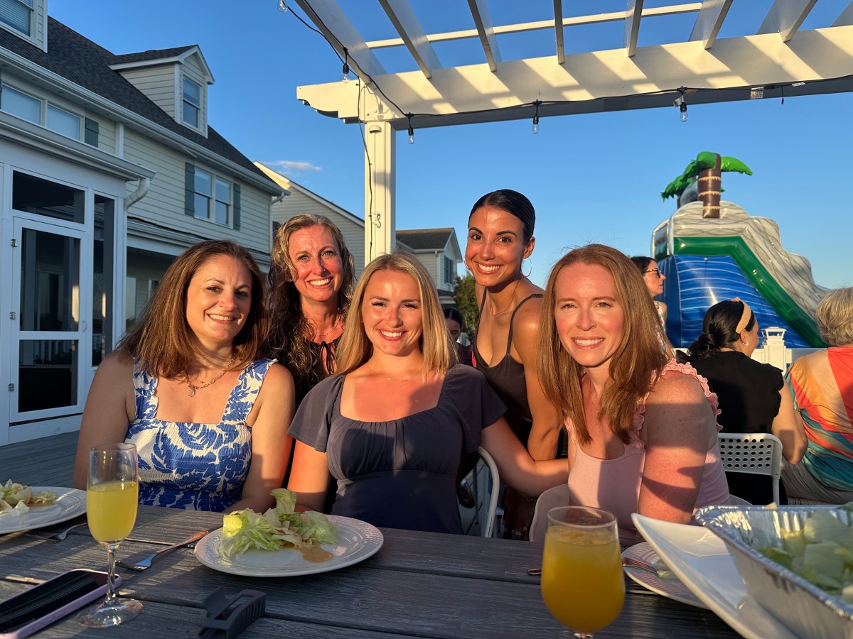Five women smiling at an outdoor gathering with food and drinks on the table.