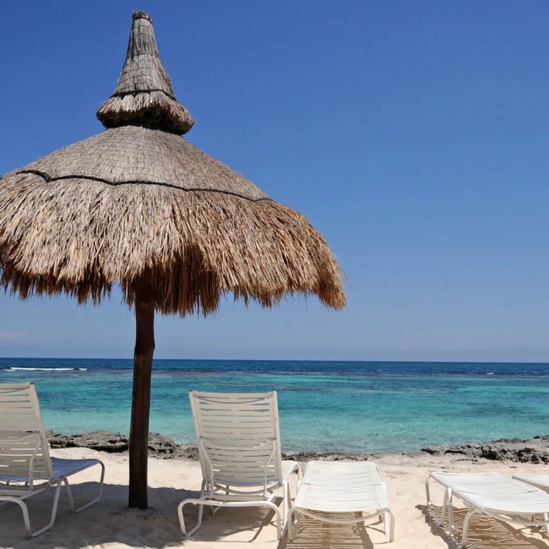Four lounge chairs under a thatched parasol on a sandy beach by the ocean.