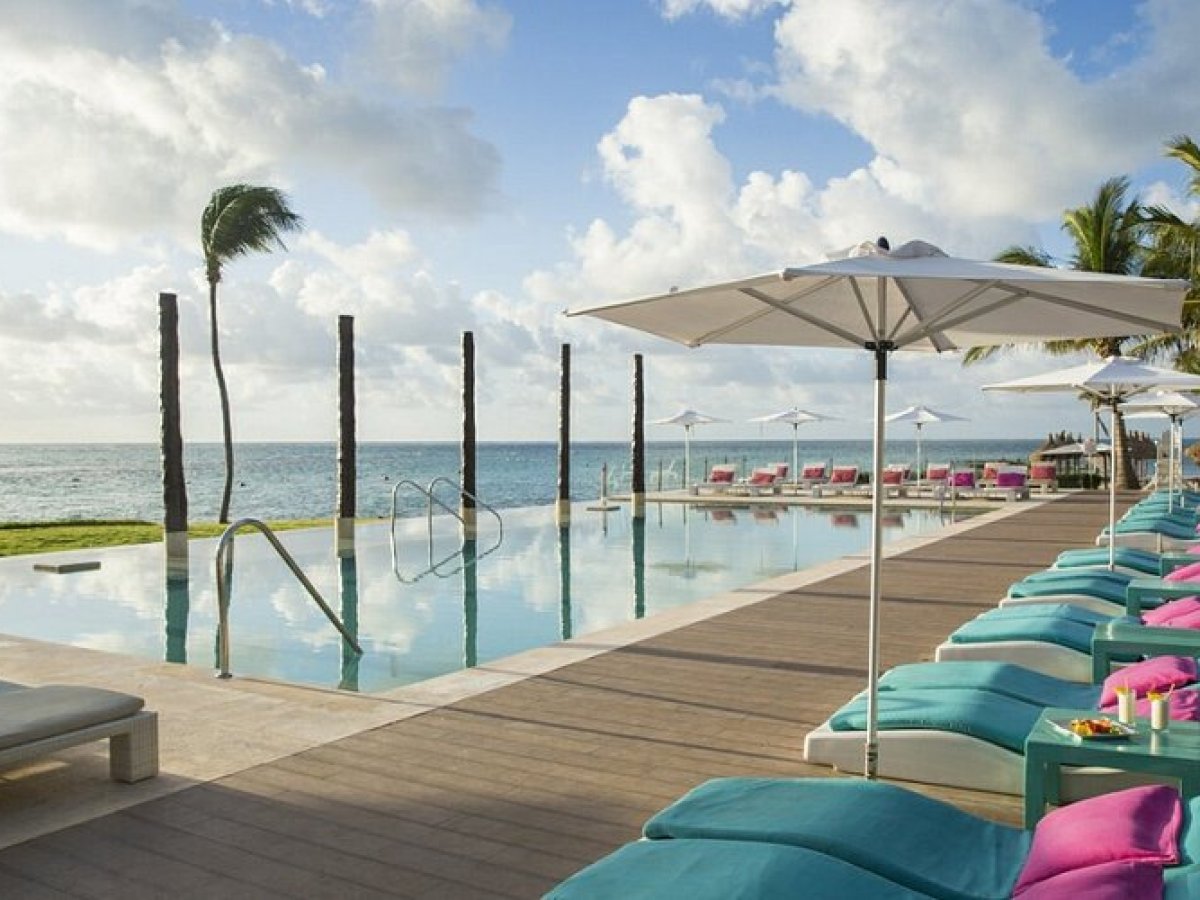 Oceanfront pool with lounge chairs, umbrellas, and palm trees under a partly cloudy sky.