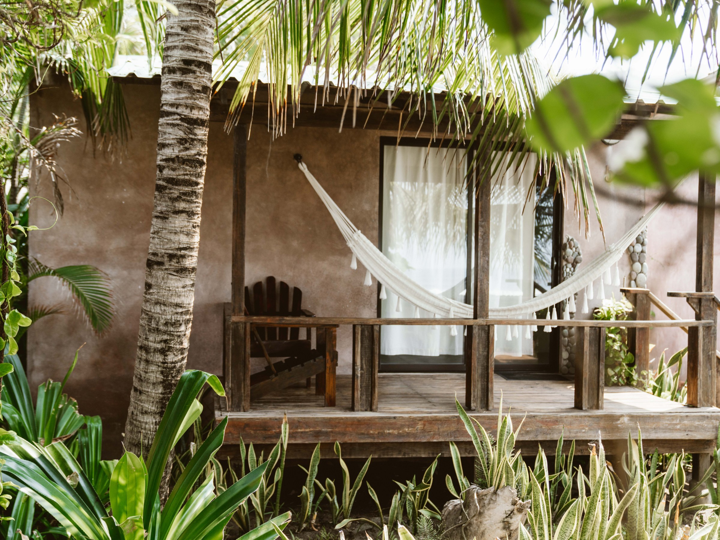 Tropical hut porch with hammock, wooden chair, and lush greenery.