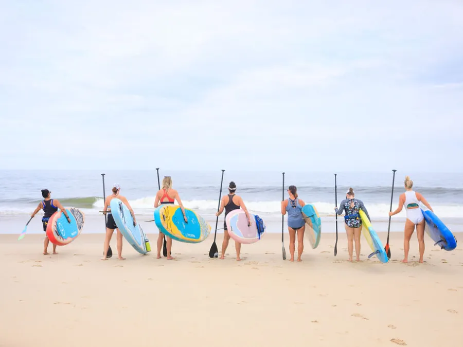 Seven people holding paddleboards face the ocean on a sandy beach.