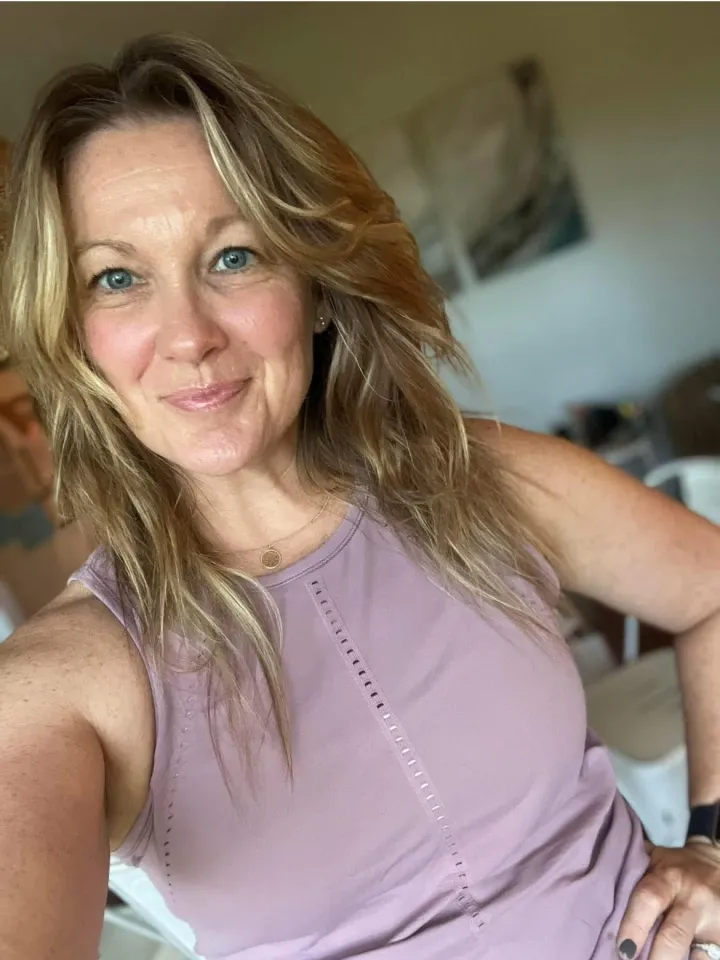 Person with long hair smiling, wearing a sleeveless purple top in an indoor setting.