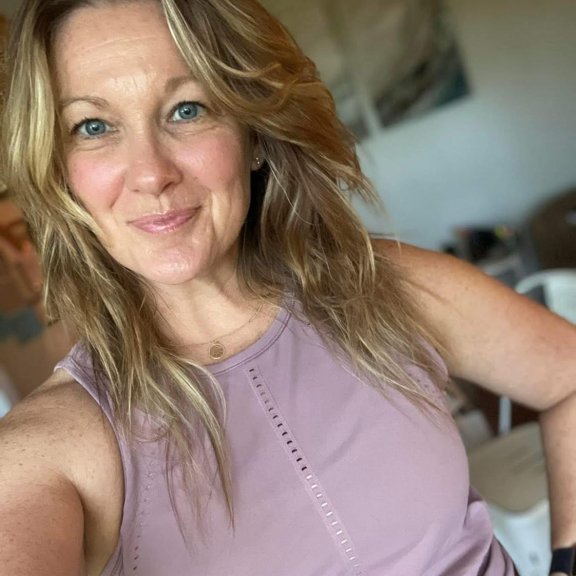 Person with long hair smiling, wearing a sleeveless purple top in an indoor setting.