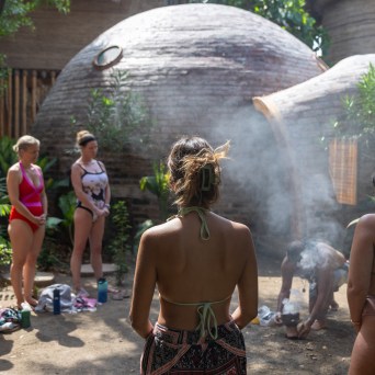 Group of people in swimsuits standing in a circle near domed structures and smoke outdoors.