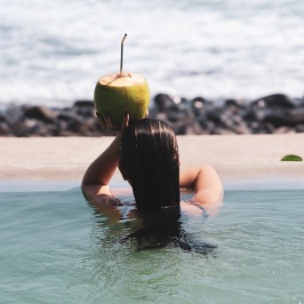 Woman in a pool holding a coconut with a straw, ocean in the background.