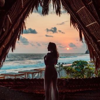Silhouette of a person standing under a thatched roof, overlooking a beach sunset.