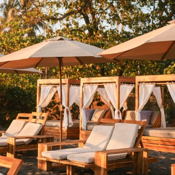 Beach lounge chairs and umbrellas on sand, surrounded by trees.