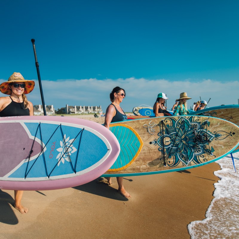 Group of people carrying colorful paddleboards on a sunny beach.