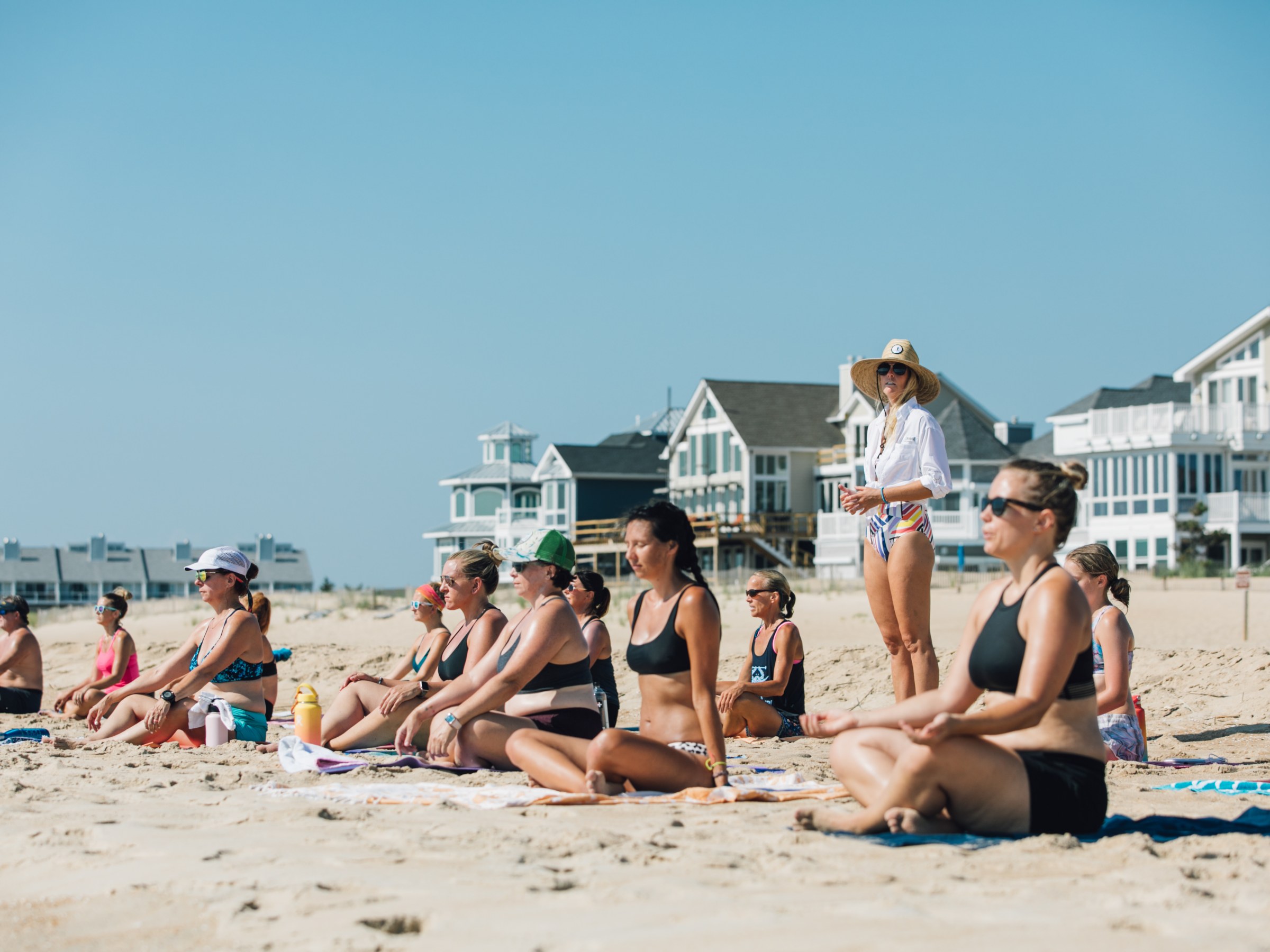Group meditating on a sunny beach with houses in the background.