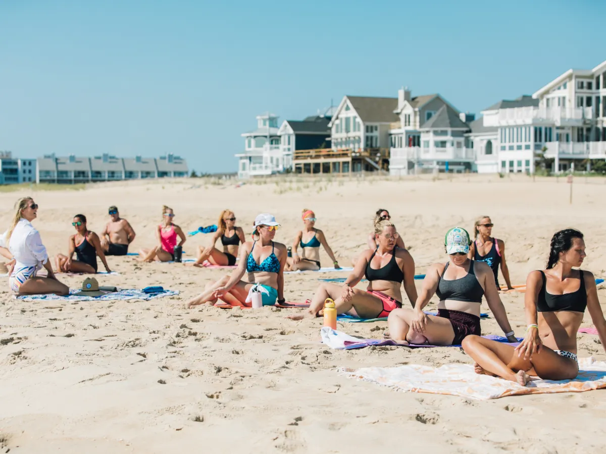 Group of people doing yoga on a beach with houses in the background.
