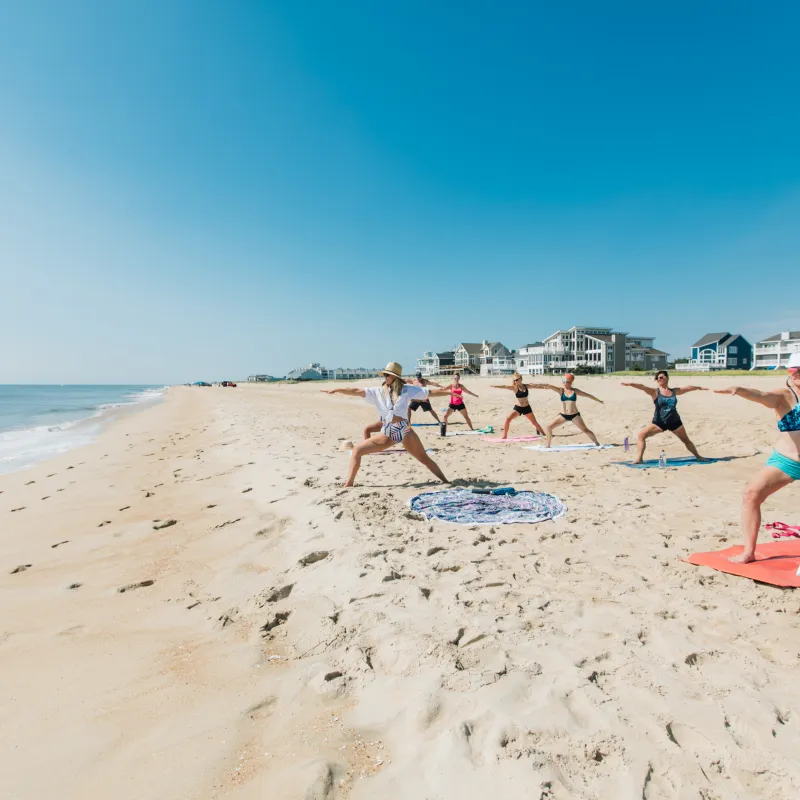 Group doing yoga on a sunny beach with ocean and houses in the background.