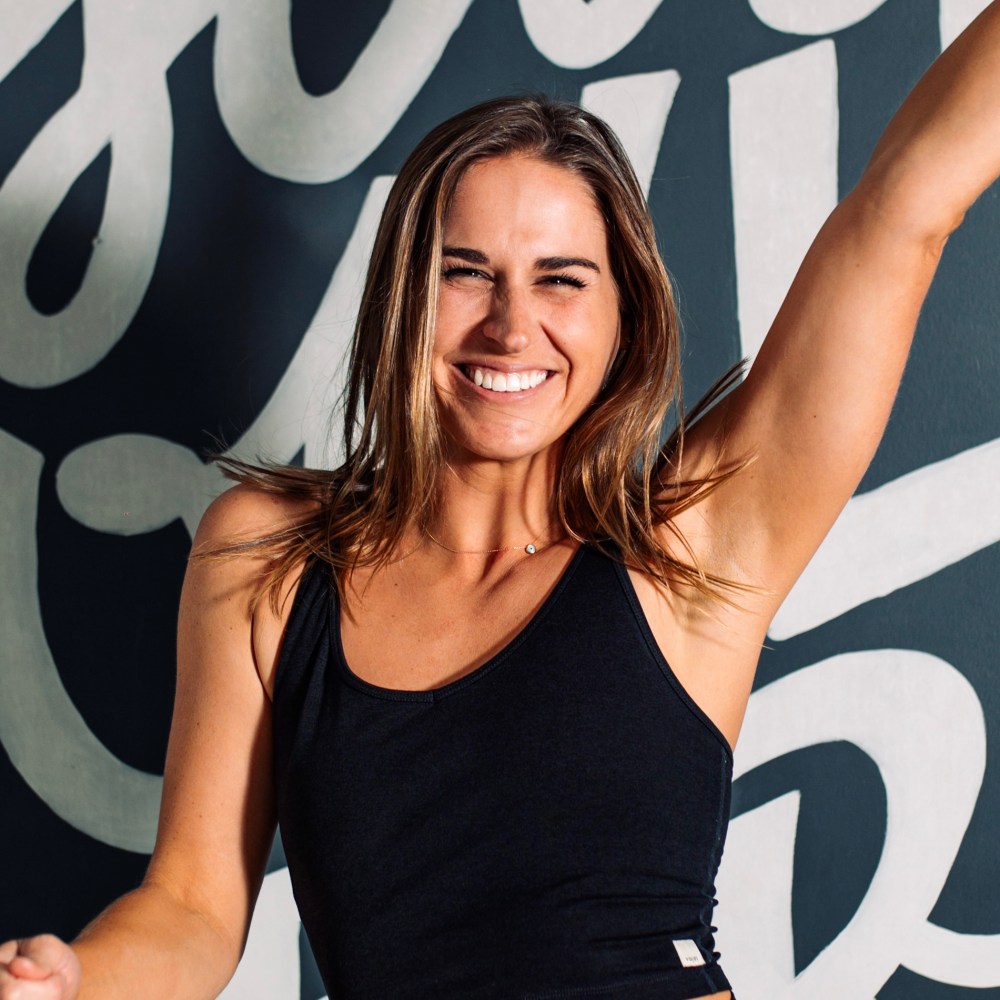 Smiling woman with arm raised, wearing a black top, against a patterned background.