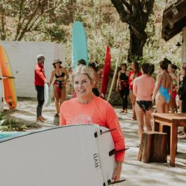 Woman in pink shirt holding a surfboard, with people and surfboards in background.
