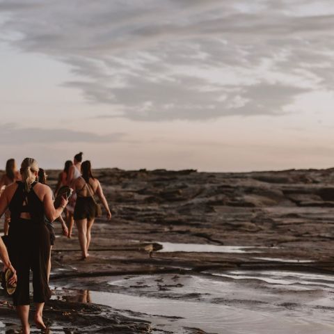 People walking on rocky terrain near water under a cloudy sky at sunset.